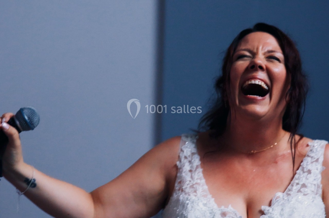 Femme souriante en robe blanche tenant un microphone, exprimant de la joie dans un environnement intérieur.