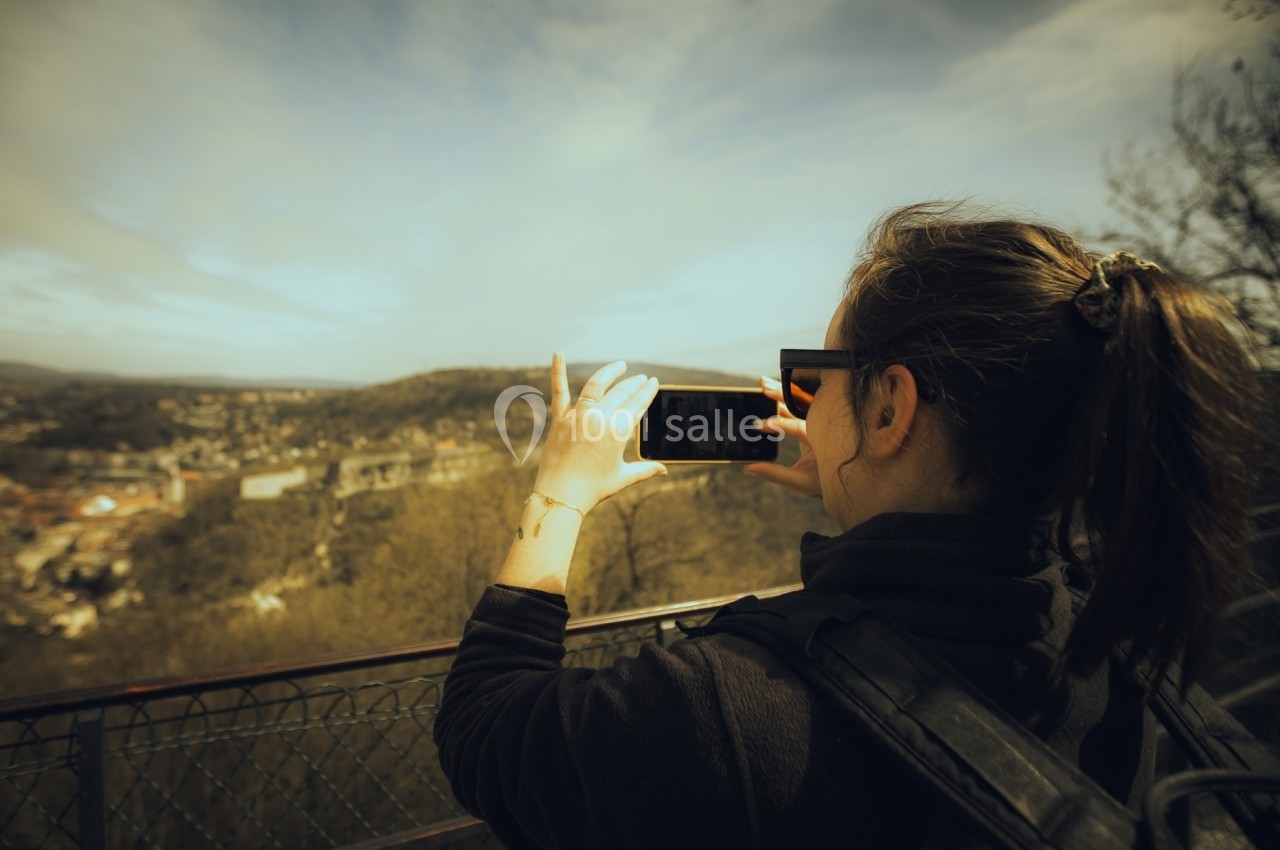 Une femme photographie un paysage urbain depuis un point de vue surélevé, sous un ciel partiellement nuageux.