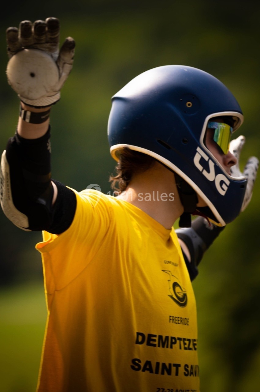 Personne portant un casque et des gants, levant les bras en extérieur, avec un fond flou de végétation.