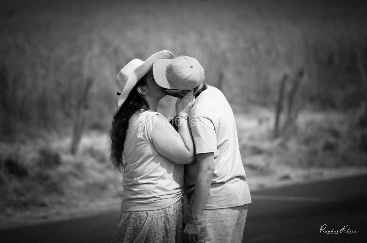 Un couple s'embrasse tendrement en extérieur, dans un paysage rural avec un champ flou en arrière-plan.