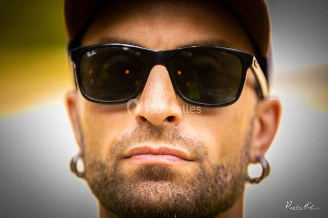 Homme portant des lunettes de soleil noires et une casquette, vu de près, avec un arrière-plan flou.