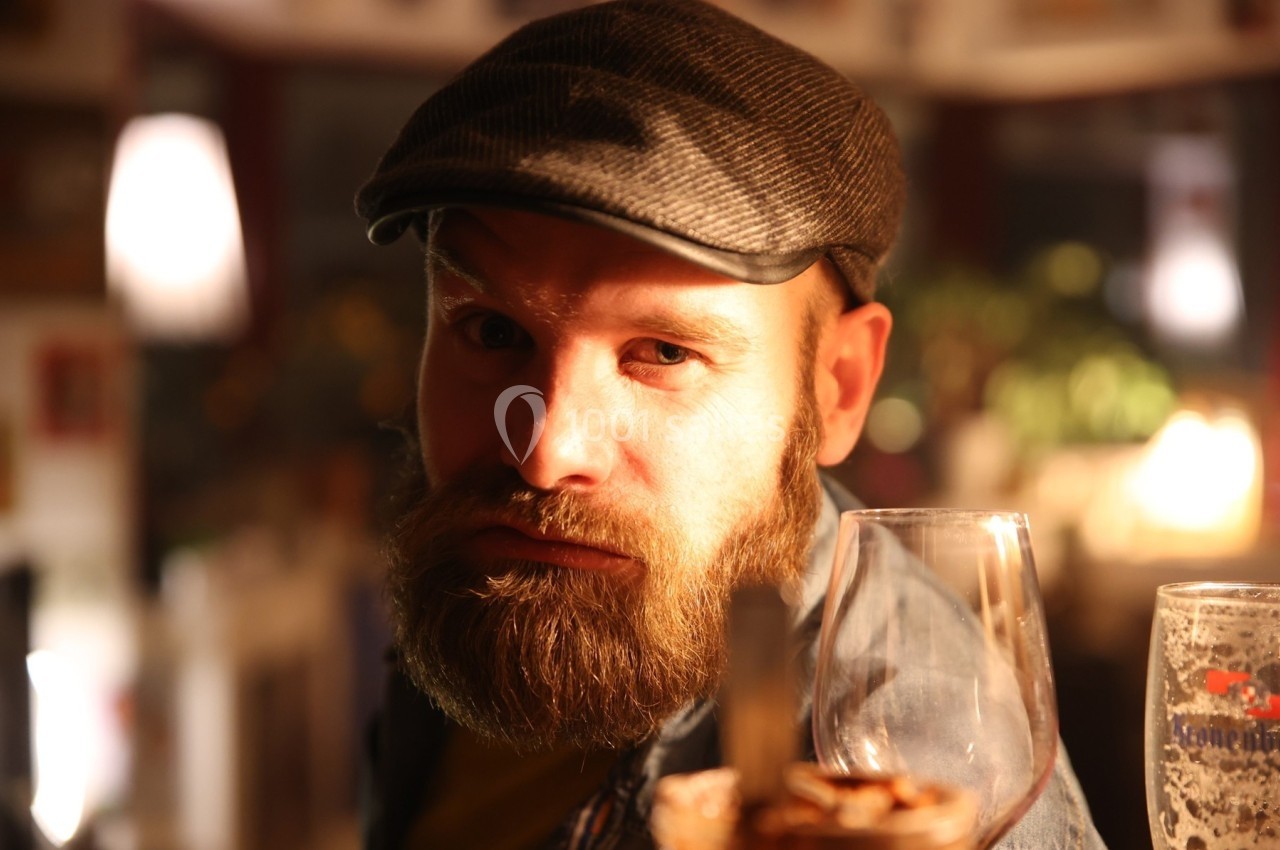 Homme barbu portant une casquette, assis dans un lieu éclairé avec des verres au premier plan.