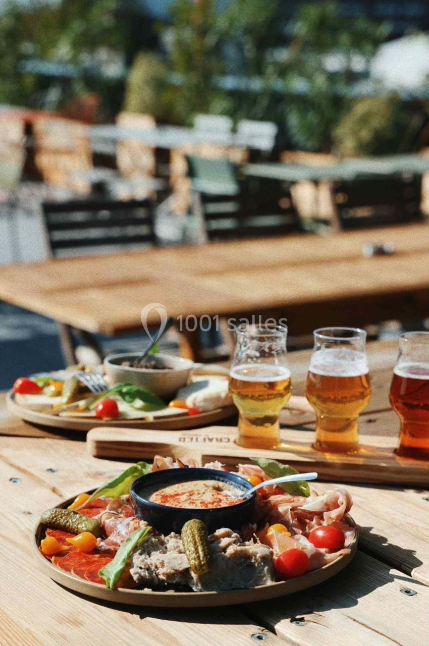 Plateau de charcuterie et légumes avec une sauce au centre, accompagné de bières sur une table en terrasse ensoleillée.