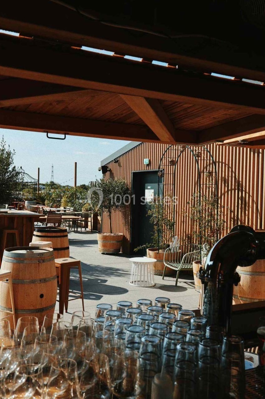 Terrasse ensoleillée avec tables, chaises, tonneaux en bois et plantes, vue depuis un comptoir avec verres empilés.