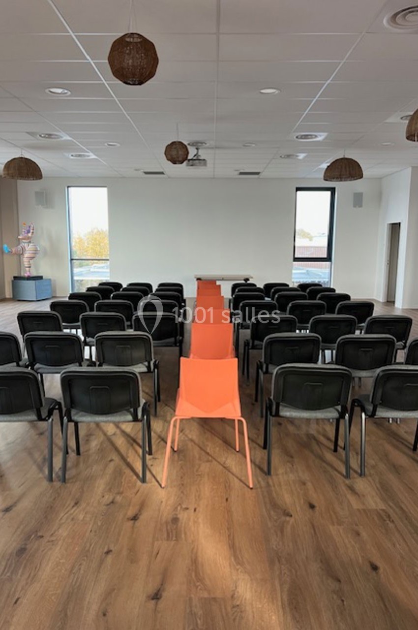 Salle de conférence avec des rangées de chaises noires et une rangée centrale de chaises orange sur un sol en bois.