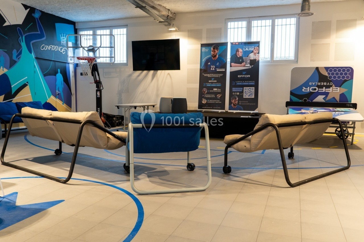 Salle aménagée avec des canapés, un écran mural, un panier de basket et des affiches promotionnelles.