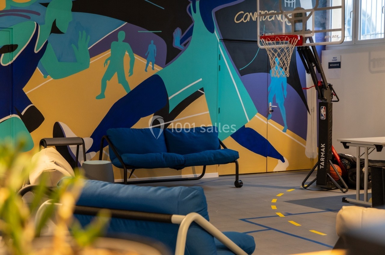 Salle colorée avec un mur illustré, un panier de basketball, des canapés bleus et un sol marqué de lignes jaunes.