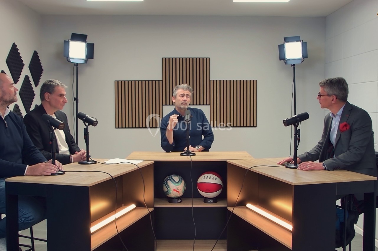 Quatre hommes discutent autour de tables équipées de micros dans un studio avec éclairage et panneaux acoustiques.
