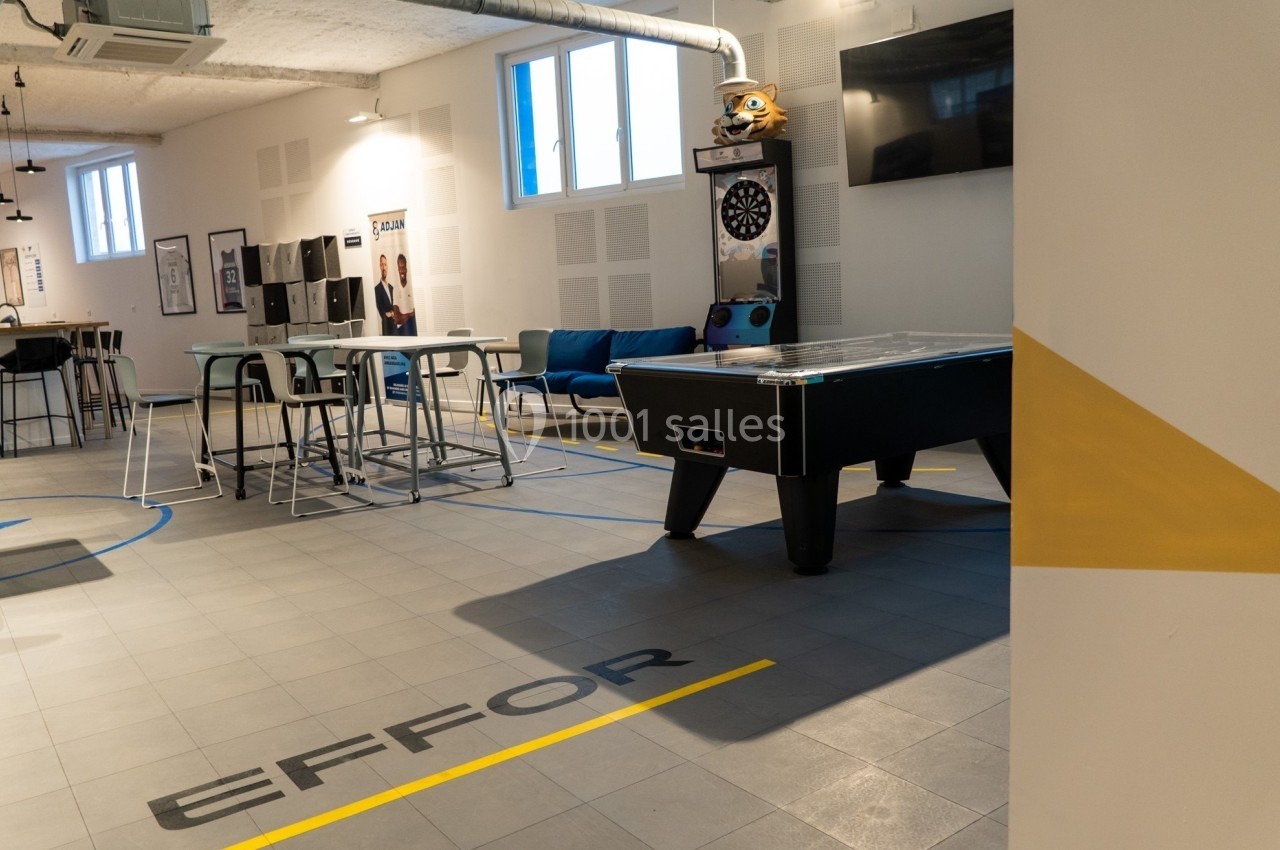 Salle de détente avec baby-foot, fléchettes, tables hautes et canapés dans un espace lumineux et moderne.
