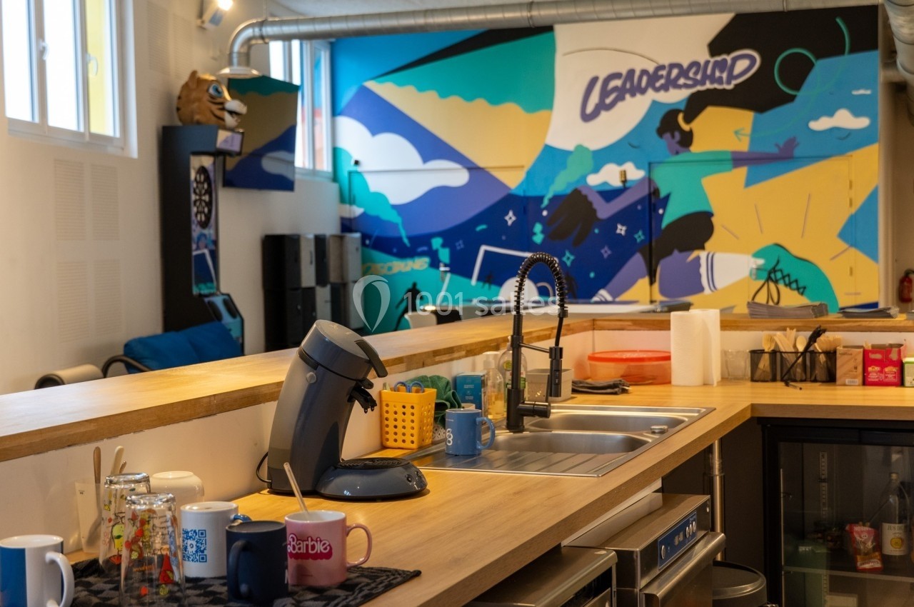 Cuisine partagée avec un comptoir en bois, évier, machine à café, mugs et mur décoré d'une fresque colorée.