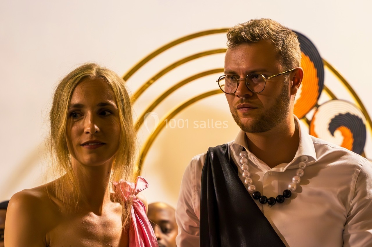 Un homme et une femme debout, vêtus de tenues élégantes, devant un fond décoratif avec des arcs dorés.