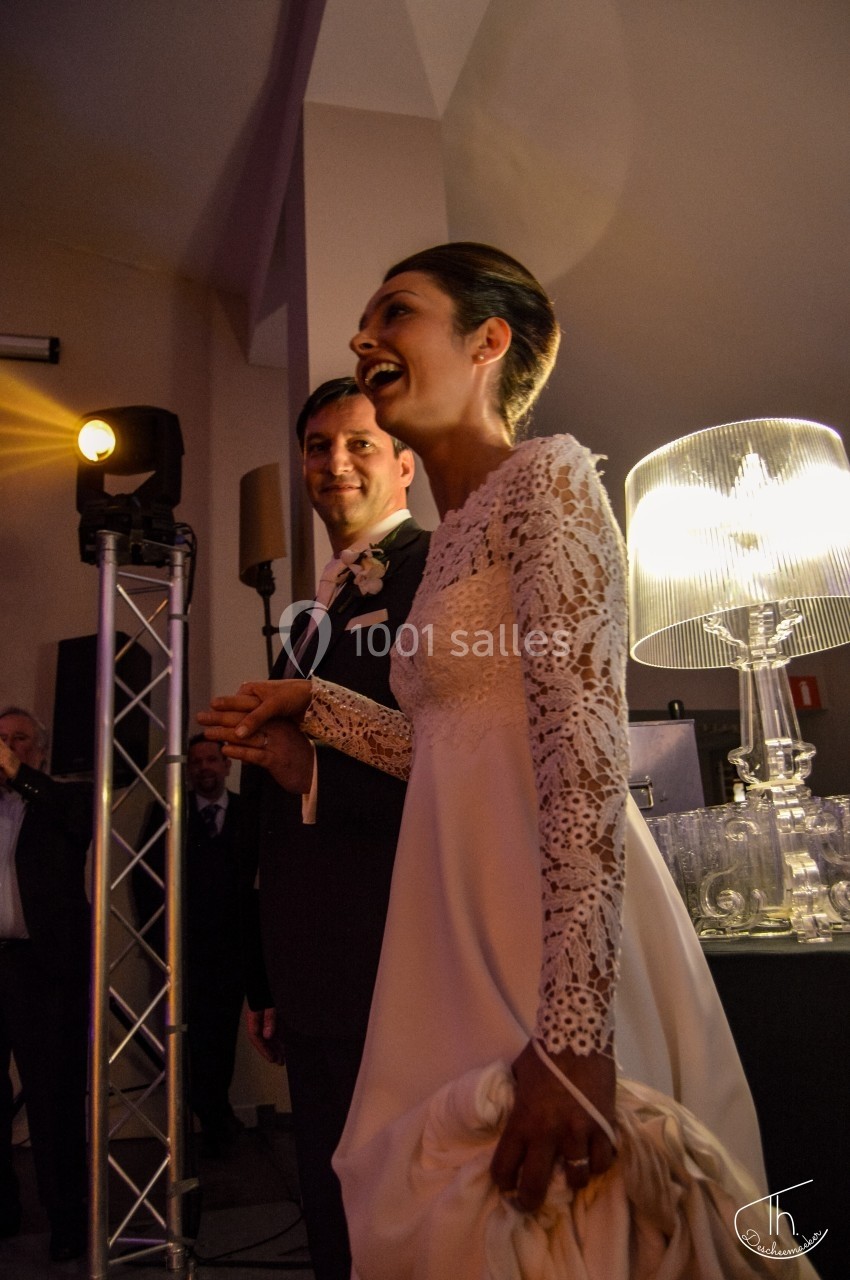 Un couple en tenue de mariage sourit lors d'une réception, éclairée par une lampe et des projecteurs.