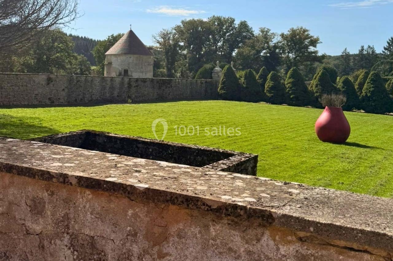 Vue d'un jardin avec pelouse, sculpture rouge en forme de vase, mur en pierre et tourelle en arrière-plan.