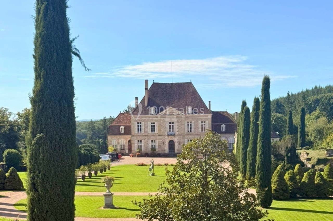 Manoir entouré de jardins verdoyants avec allées bordées de cyprès sous un ciel bleu clair.