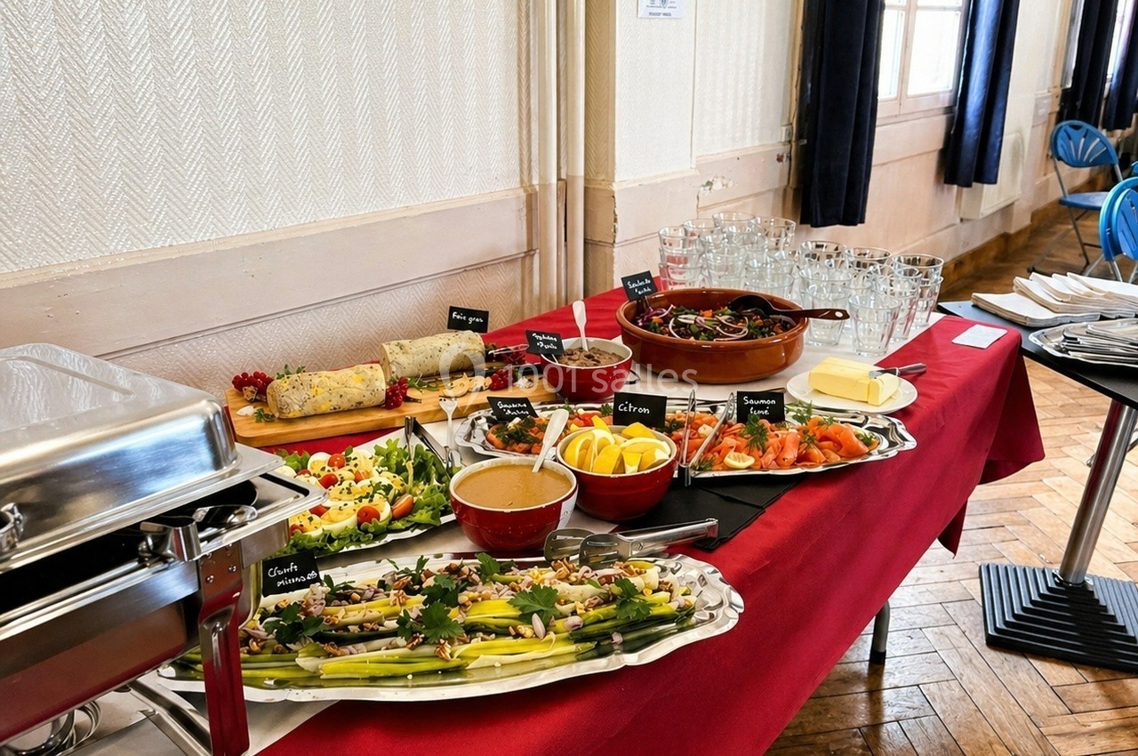 Buffet varié avec plats froids, salades, légumes, saumon fumé, sauces et verres disposés sur une table rouge.