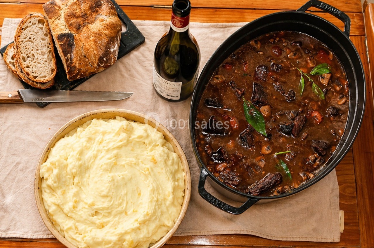 Plat de bœuf mijoté avec légumes, purée de pommes de terre, pain artisanal et bouteille de vin sur une table en bois.