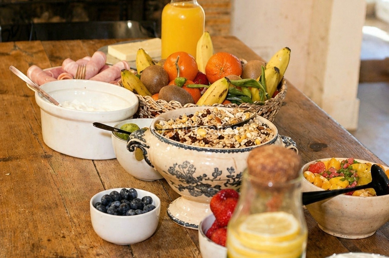 Petit-déjeuner avec fruits frais, muesli, yaourt, jus d'orange et charcuterie sur une table en bois.