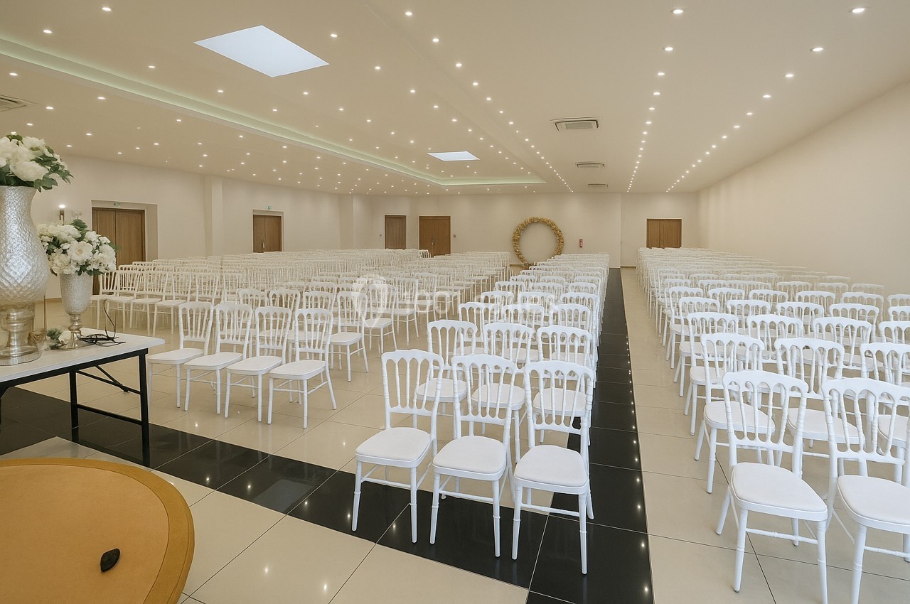 Salle de réception lumineuse avec des rangées de chaises blanches alignées et une arche décorative au fond.