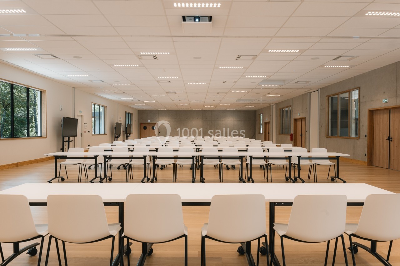 Salle de conférence vide avec des rangées de tables et de chaises blanches, éclairée par des lumières au plafond.