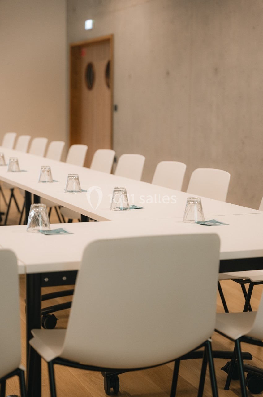 Salle de réunion avec une grande table blanche en angle, des chaises blanches et des verres posés sur des sets de table.