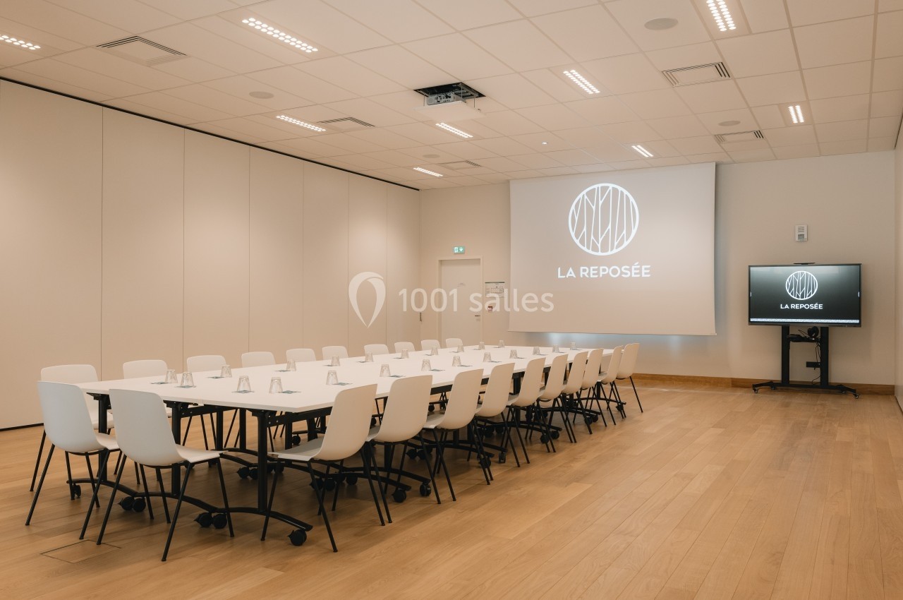 Salle de réunion lumineuse avec une grande table rectangulaire, des chaises blanches et un écran de projection allumé.