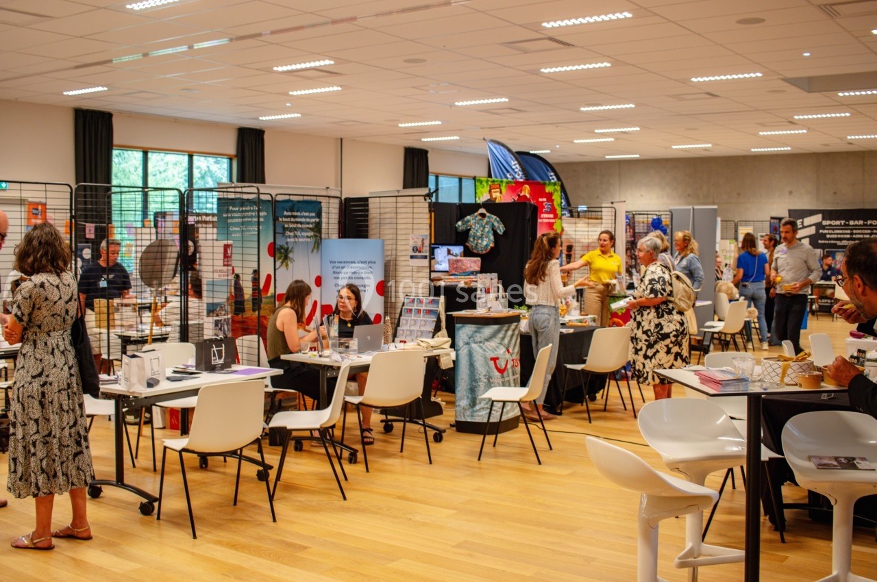 Salle d'exposition avec stands d'information, visiteurs échangeant et explorant divers produits et services.