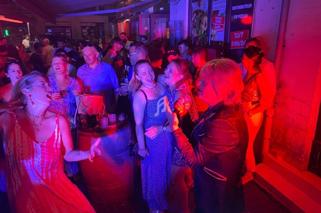 Groupe de personnes dans un bar animé, éclairé par des lumières rouges, certaines chantant et dansant.