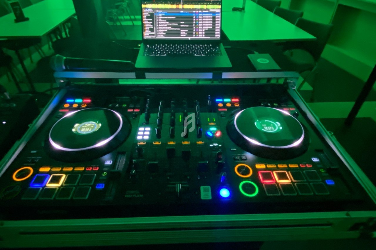 Table de mixage DJ éclairée avec des lumières colorées, un ordinateur portable affichant un logiciel audio en arrière-plan.