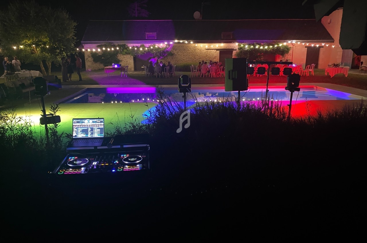 Console de DJ en premier plan face à une piscine éclairée de lumières colorées lors d'une soirée en extérieur.
