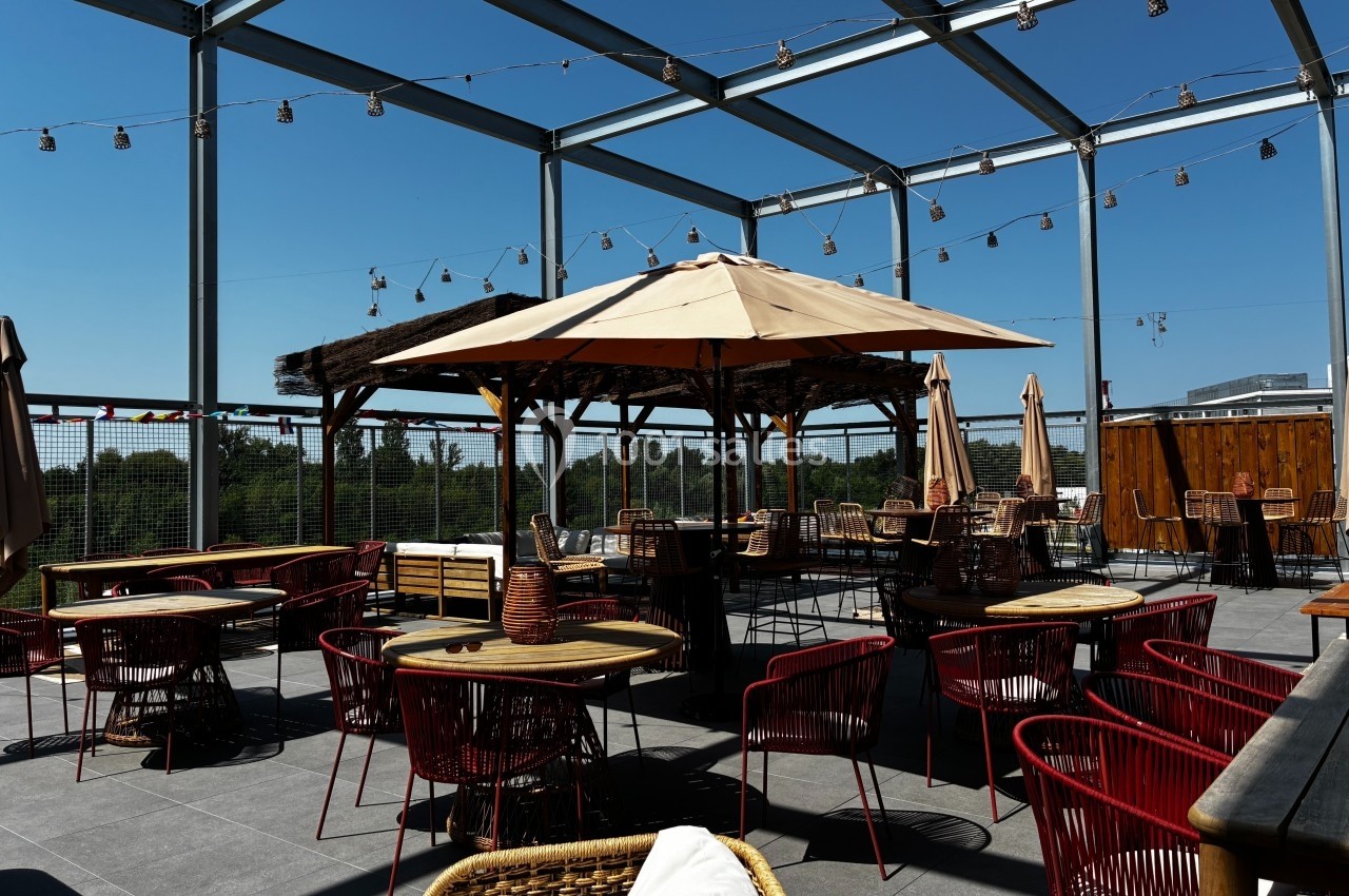 Terrasse ensoleillée avec tables, chaises rouges et parasols, entourée de guirlandes lumineuses et d'une structure…