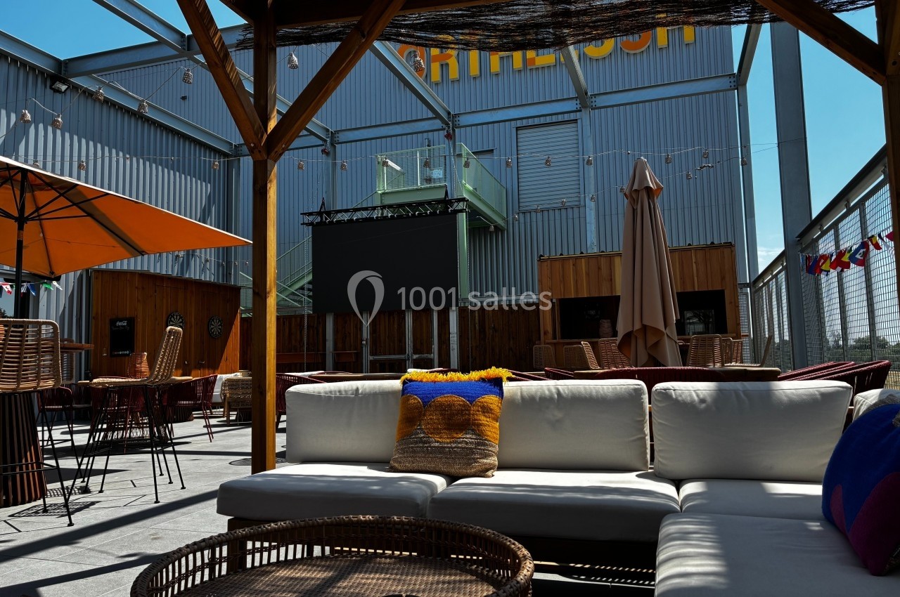 Espace extérieur avec canapés, parasols et écran géant, entouré de structures métalliques et de bois.