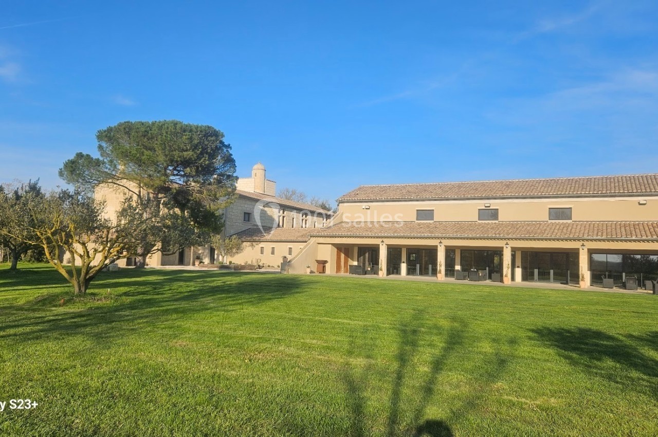 Grande maison en pierre avec toit en tuiles, entourée d'une pelouse et d'arbres sous un ciel bleu clair.