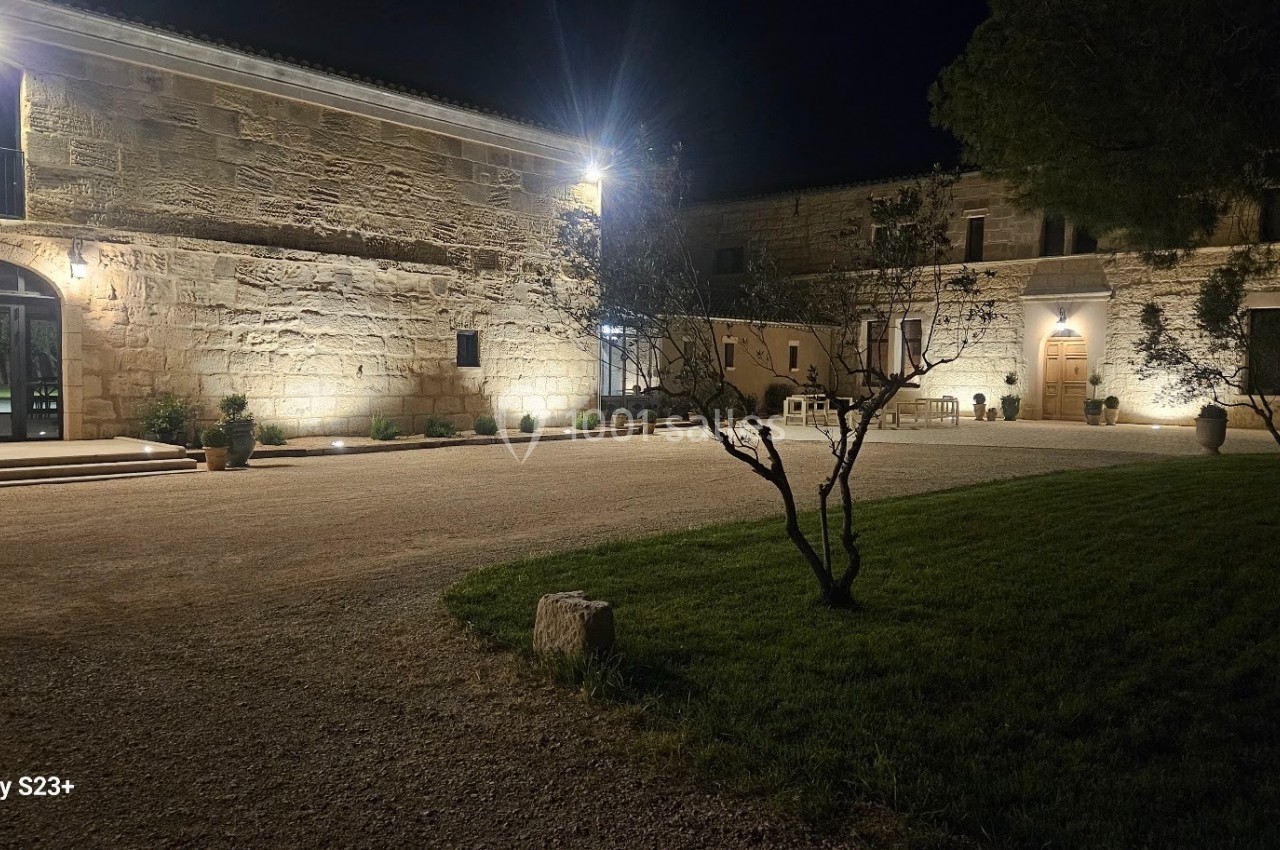 Cour d'une grande bâtisse en pierre éclairée la nuit, avec des arbres et un espace gazonné au premier plan.