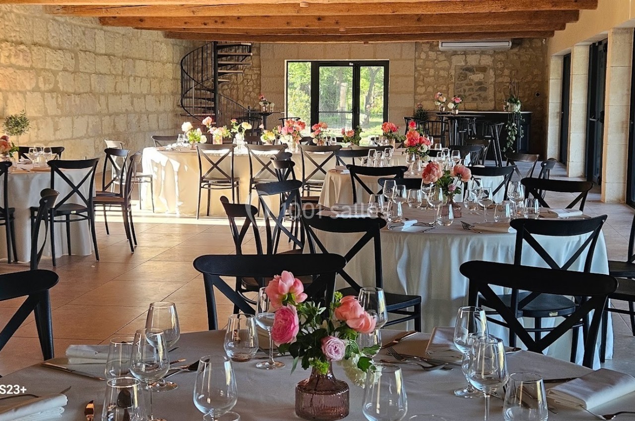 Salle de réception lumineuse avec tables rondes décorées de fleurs et chaises noires, vue sur un escalier en colimaçon.