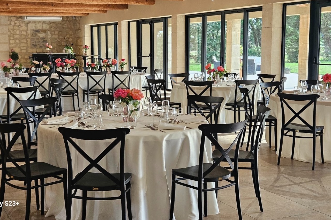 Salle de réception lumineuse avec tables rondes dressées, nappes blanches et bouquets de fleurs colorées.