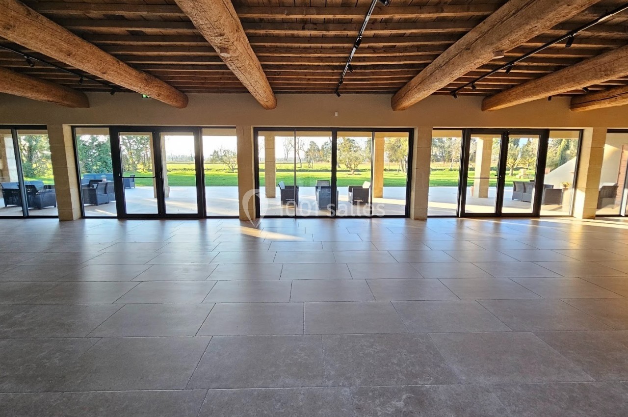 Salle spacieuse avec sol en carrelage, plafond en poutres apparentes et grandes baies vitrées donnant sur un espace vert.