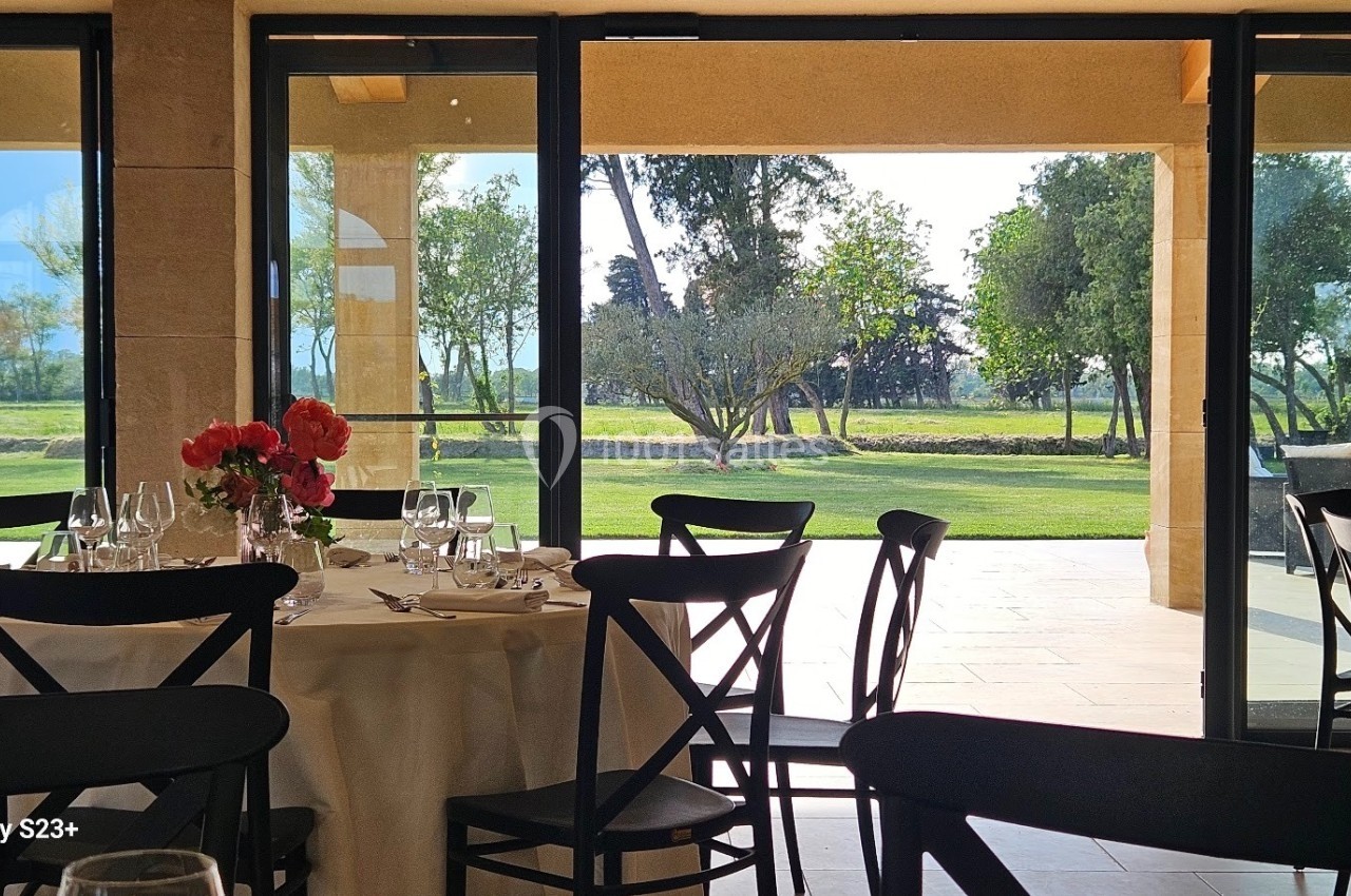 Salle de restaurant lumineuse avec vue sur un jardin verdoyant à travers de grandes baies vitrées.