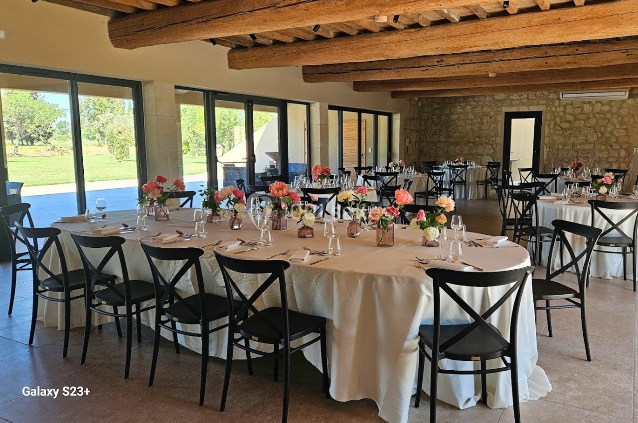 Salle de réception lumineuse avec tables rondes décorées de fleurs colorées, chaises noires et grandes baies vitrées.