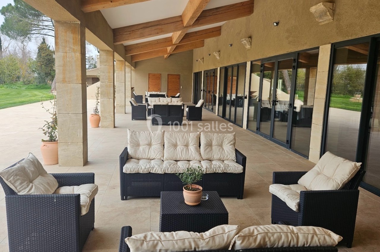 Terrasse couverte avec mobilier en rotin noir et coussins blancs, entourée de baies vitrées et donnant sur un jardin.