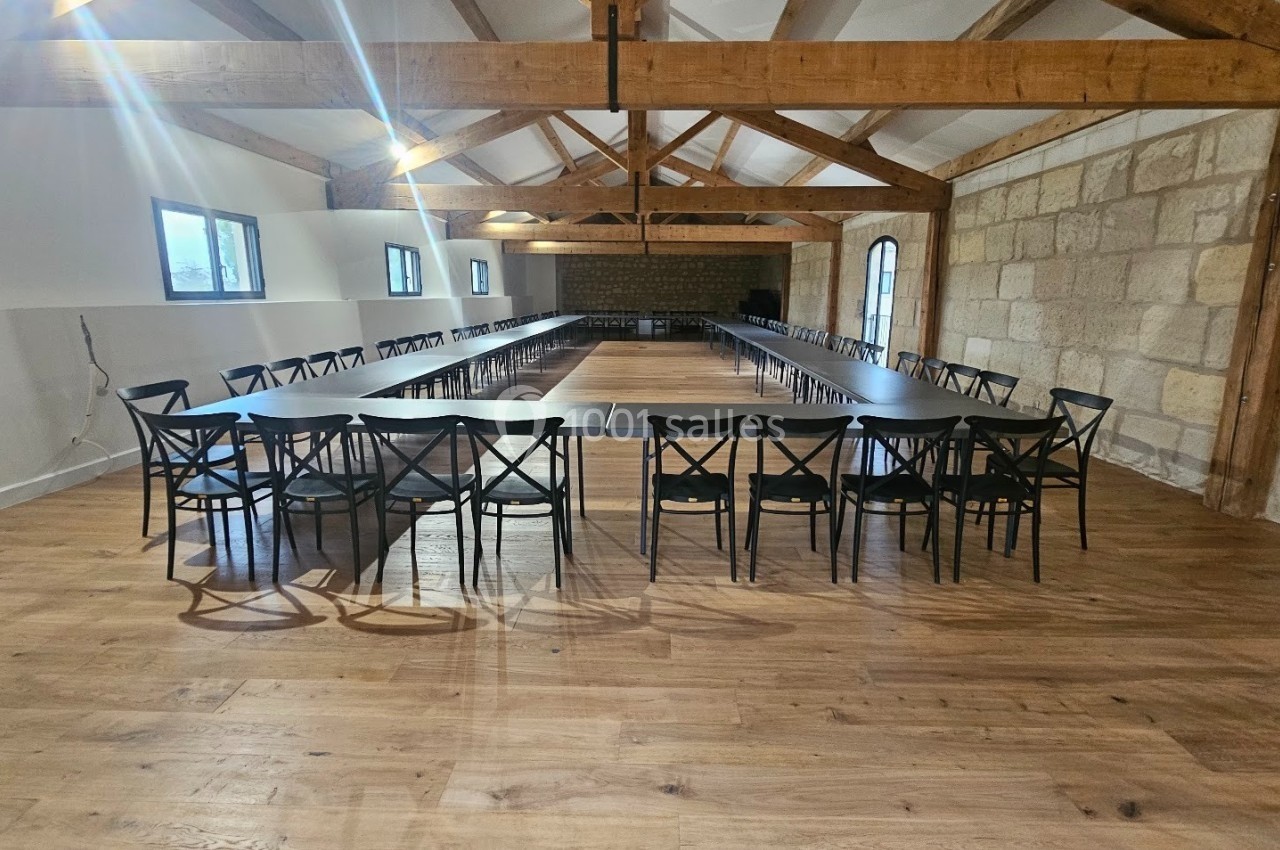 Salle lumineuse avec poutres apparentes, tables disposées en U et chaises noires sur un sol en parquet.