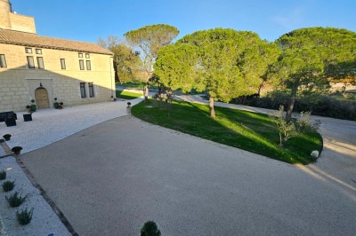 Bâtisse en pierre avec une verrière, entourée d'une cour pavée et d'espaces verts sous un ciel dégagé.