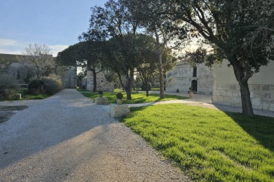 Bâtisse en pierre avec une verrière, entourée d'une cour pavée et d'espaces verts sous un ciel dégagé.