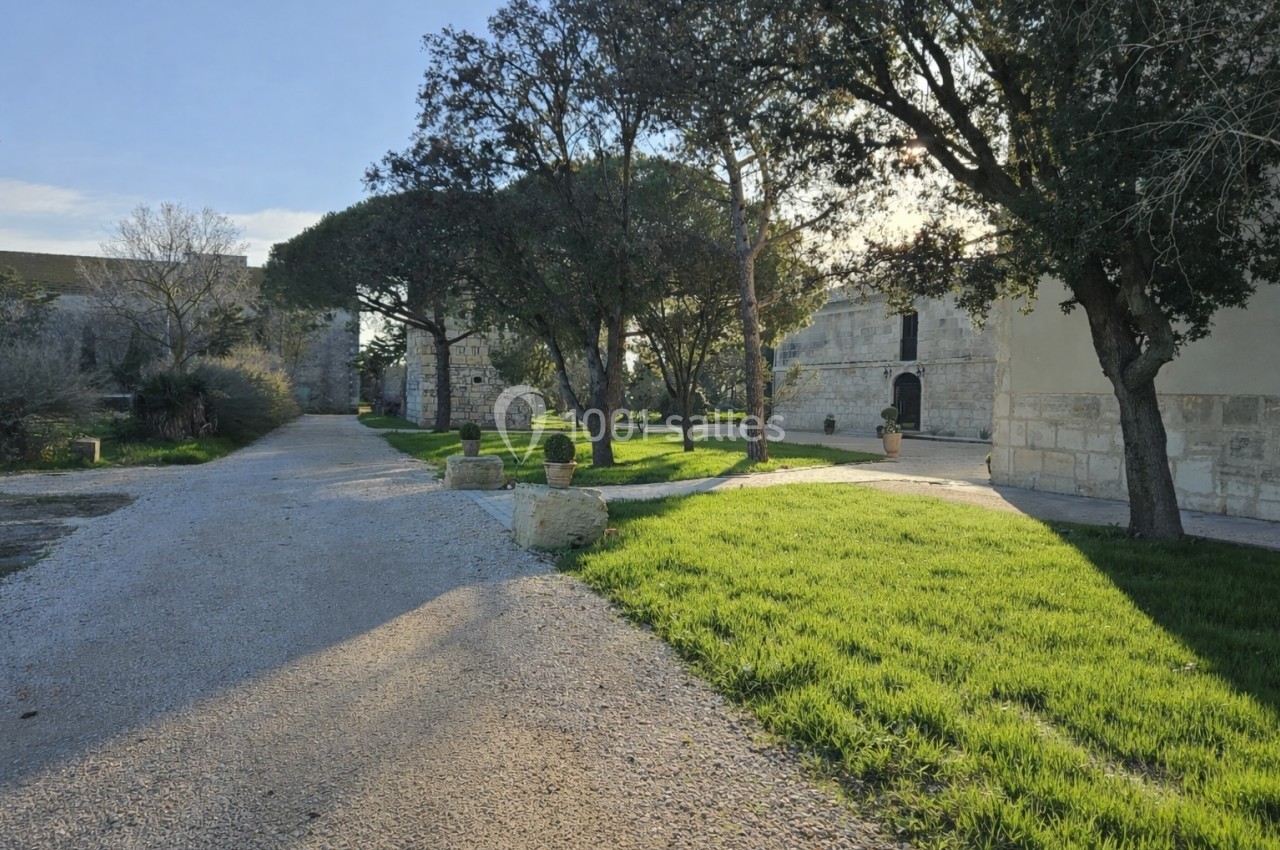Allée bordée d'arbres et de pelouses, menant à des bâtiments en pierre sous un ciel dégagé.