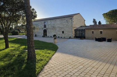 Bâtisse en pierre avec une verrière, entourée d'une cour pavée et d'espaces verts sous un ciel dégagé.