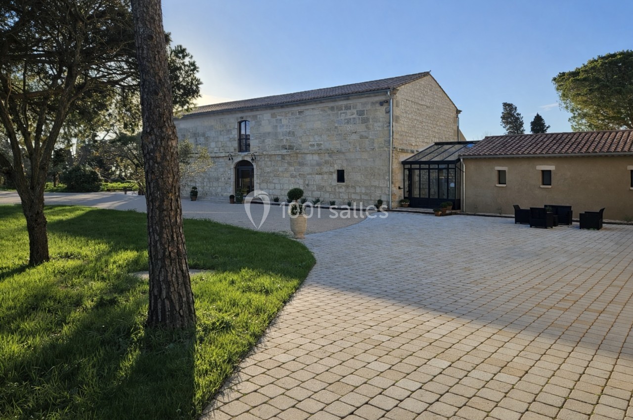 Bâtisse en pierre avec une verrière, entourée d'une cour pavée et d'espaces verts sous un ciel dégagé.