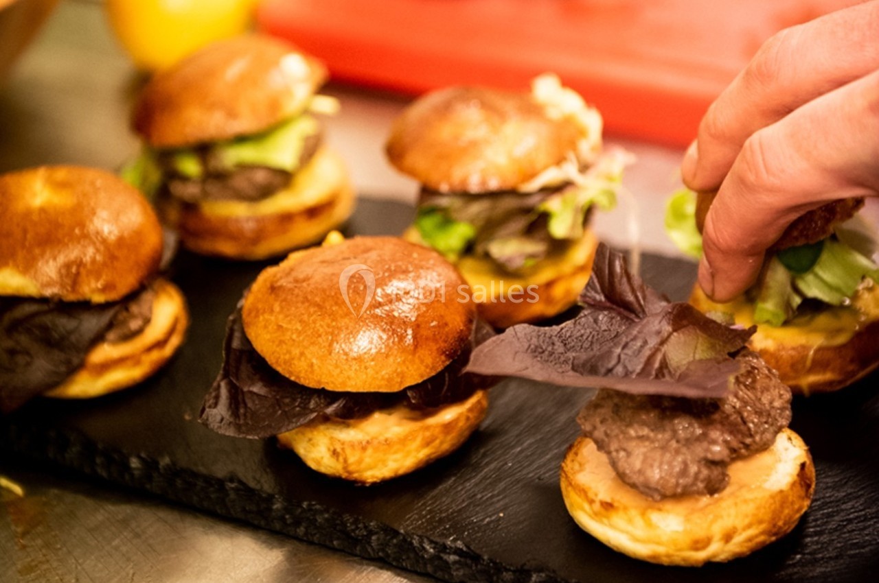 Mini-burgers garnis de viande, salade et sauce, disposés sur une ardoise, avec une main ajustant la garniture.