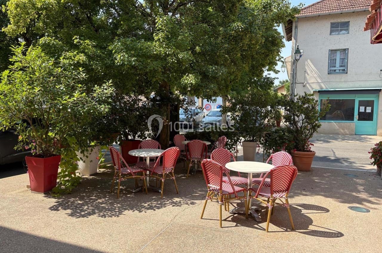 Terrasse extérieure avec tables et chaises rouges sous des arbres, entourée de plantes et de bâtiments.