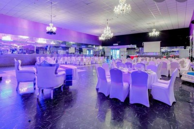 Salle de réception spacieuse avec tables dressées, chaises blanches, piste de danse et éclairage violet.