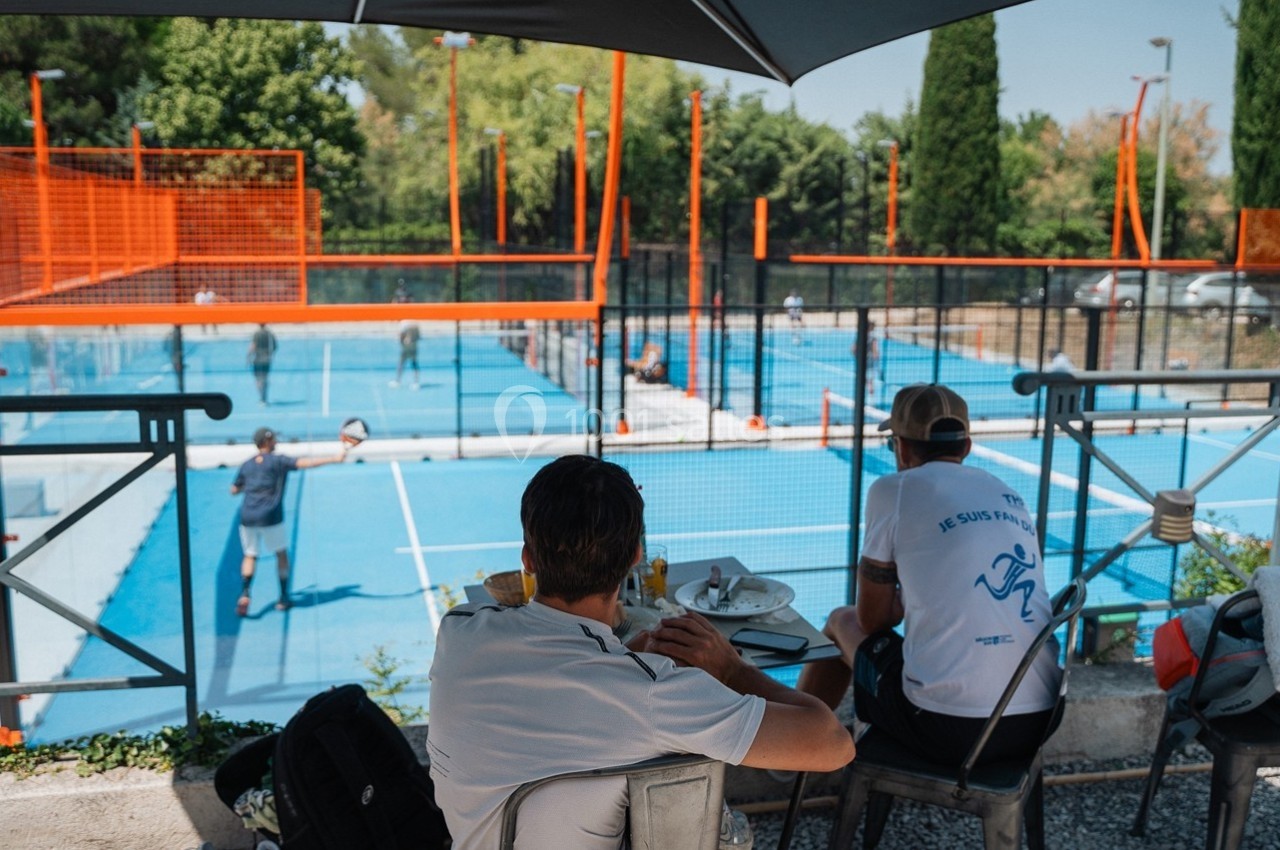 Deux hommes assis à une table en terrasse regardent des joueurs sur des terrains de padel extérieurs.