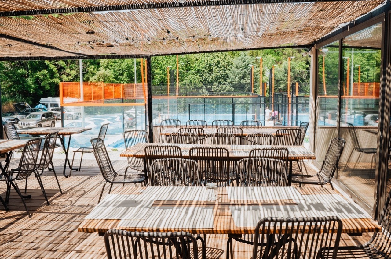 Terrasse en bois avec tables et chaises en rotin, ombragée par une pergola, vue sur une piscine et un espace arboré.